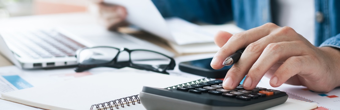 Close-up of a person using a calculator at a desk with documents, a laptop, eyeglasses and a smartphone.
